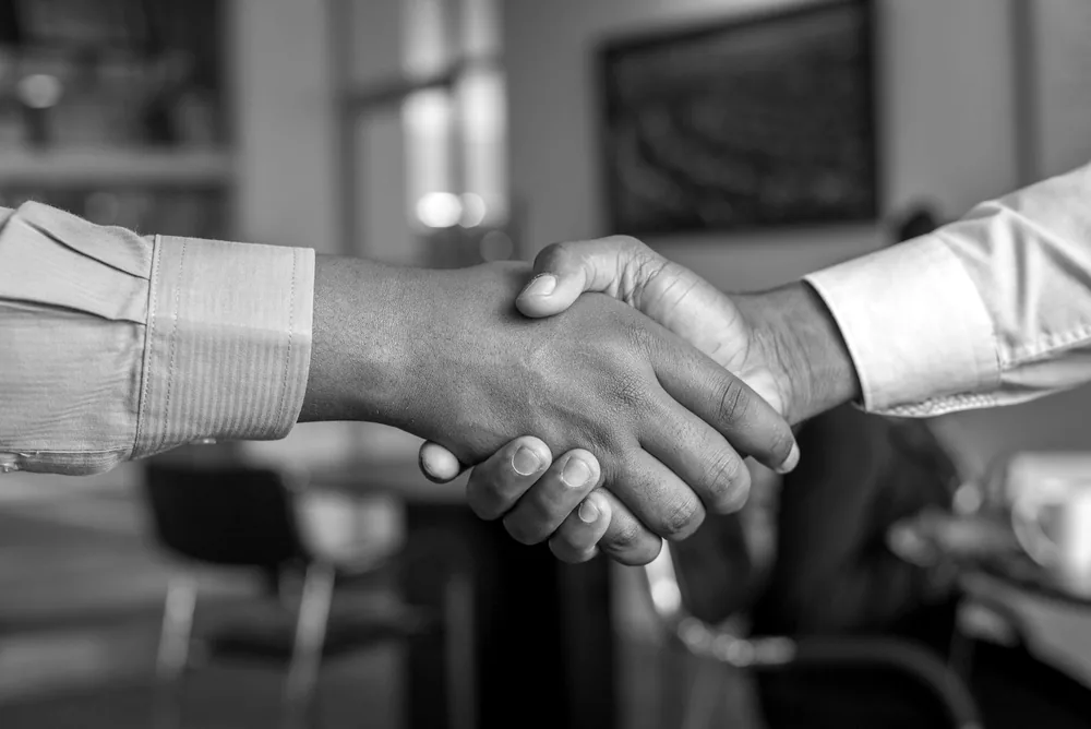 Two people shaking hands across a desk at the close of a meeting.